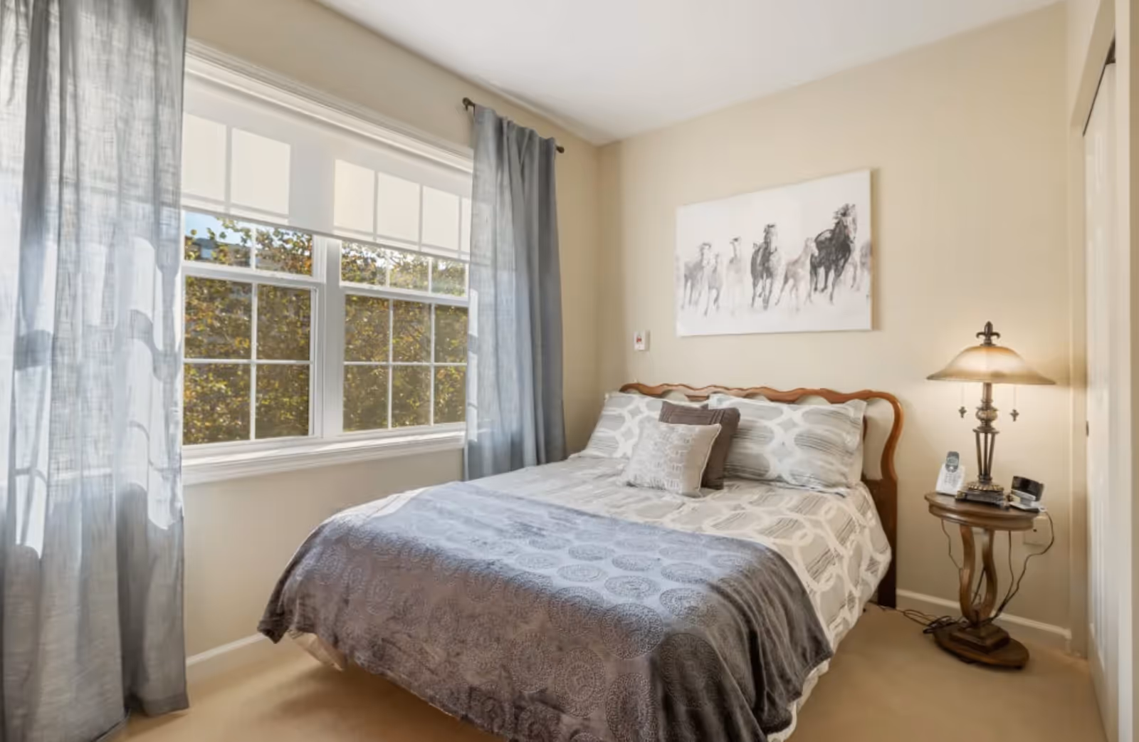 A cozy bedroom with a large window covered by sheer gray curtains and white blinds. The bed has a wooden headboard and is made with patterned gray and white bedding and decorative pillows. A small wooden side table next to the bed holds a vintage-style lamp, a cordless phone, and other small items. Above the bed hangs a painting of running horses. The walls are painted beige and the room has a warm, inviting atmosphere.