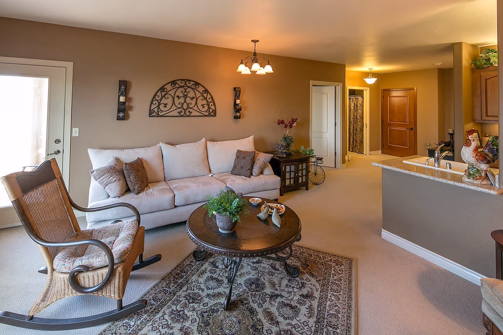 A cozy living room with a white sofa adorned with multiple cushions, a round wooden coffee table with decorative items and a potted plant, and a wicker rocking chair with a cushion. The room has beige walls with decorative wall hangings, a patterned area rug, and a view into a kitchen area with a countertop and a rooster figurine. There is a door leading outside and a hallway with doors to other rooms.