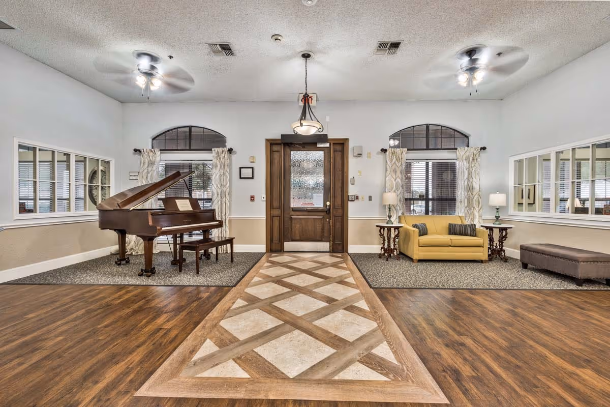 Interior view of a senior living facility common area with a wooden grand piano and bench on the left, a yellow loveseat with two striped pillows on the right, two side tables with lamps, large windows with patterned curtains, a wooden door in the center, ceiling fans with lights, and a patterned floor rug leading to the door.