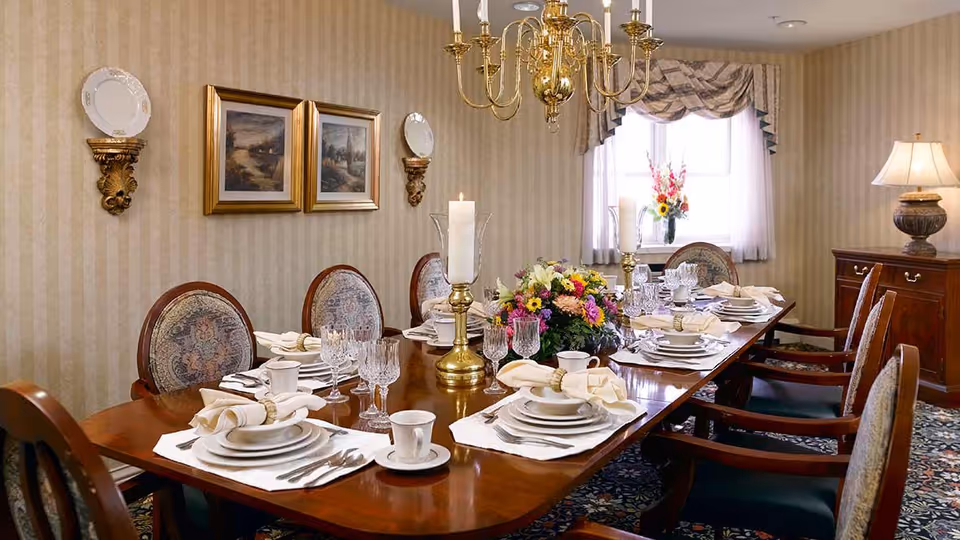 A formal dining room with a long wooden table set for a meal. The table is adorned with white plates, cups, silverware, cloth napkins with rings, crystal glasses, and two tall brass candle holders with white candles. A floral centerpiece is placed in the middle of the table. The room has patterned wallpaper, two framed paintings on the wall, decorative wall-mounted plates, a window with sheer curtains and a valance, a wooden sideboard with a lamp, and a patterned carpet.
