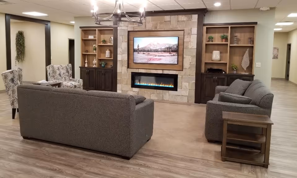 A cozy living room area in Green Gardens Assisted Living Memory Care featuring two gray sofas facing a wall-mounted TV above a modern electric fireplace. The wall is decorated with stone tiles and wooden built-in shelves on either side, holding various decorative items. Two patterned armchairs and a small wooden side table are also visible, with light wood flooring and soft lighting creating a warm atmosphere.