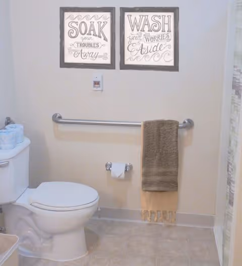 A bathroom with a white toilet, a grab bar mounted on the wall with two towels hanging on it, a toilet paper holder with a roll of toilet paper, and two framed decorative signs above the grab bar. The signs read 'SOAK your TROUBLES Away' and 'WASH your WORRIES Aside'. There are extra rolls of toilet paper on top of the toilet tank.