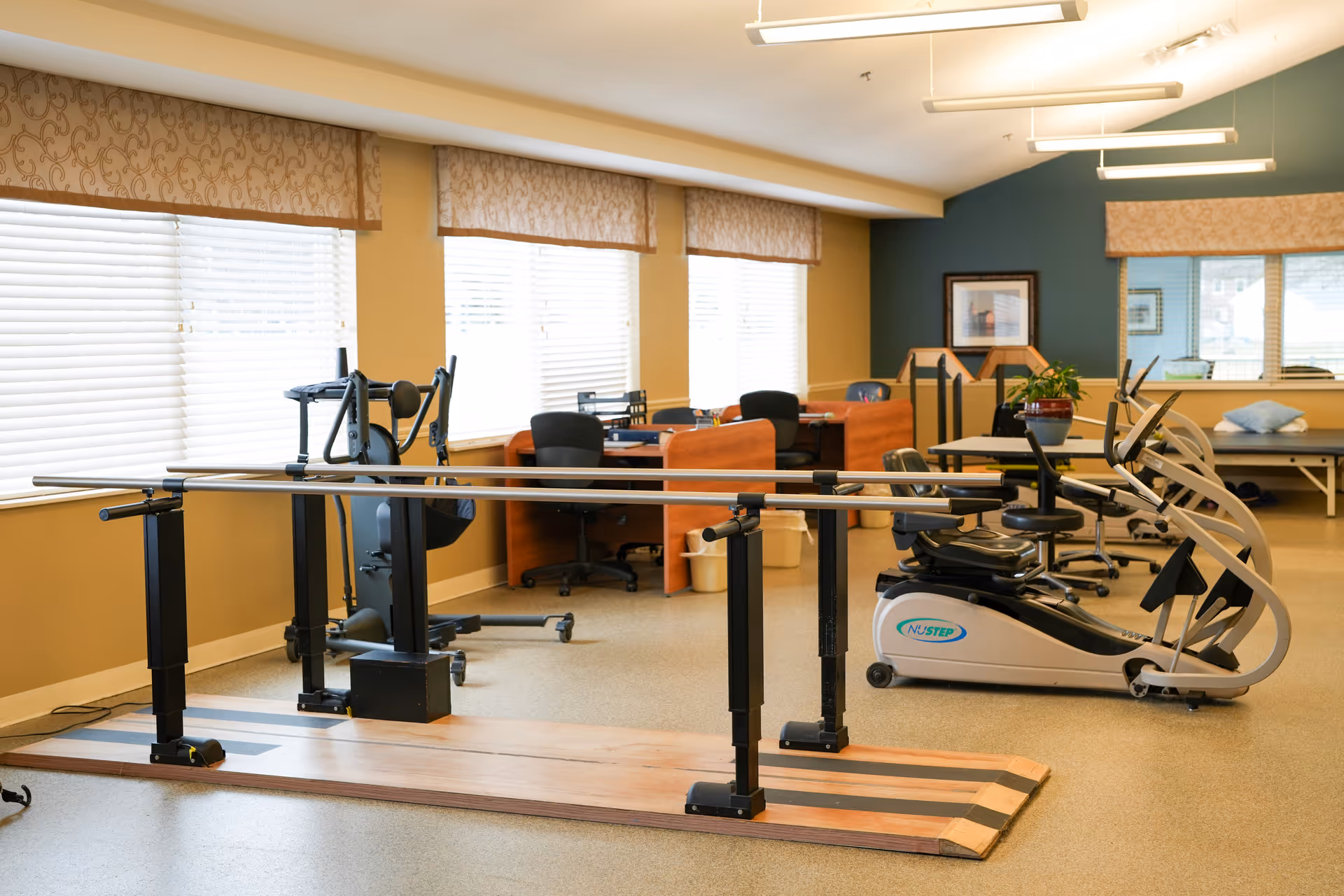 A rehabilitation or physical therapy room with parallel bars for walking exercises, exercise machines including a NuStep recumbent cross trainer, office desks with chairs, and large windows with blinds and valances letting in natural light.