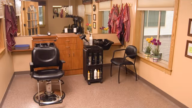 Interior of a salon area with a black salon chair in front of a wooden cabinet and a black sink. There is a black chair next to the sink, colorful scarves hanging on the wall, and a window with a flower pot on the windowsill.