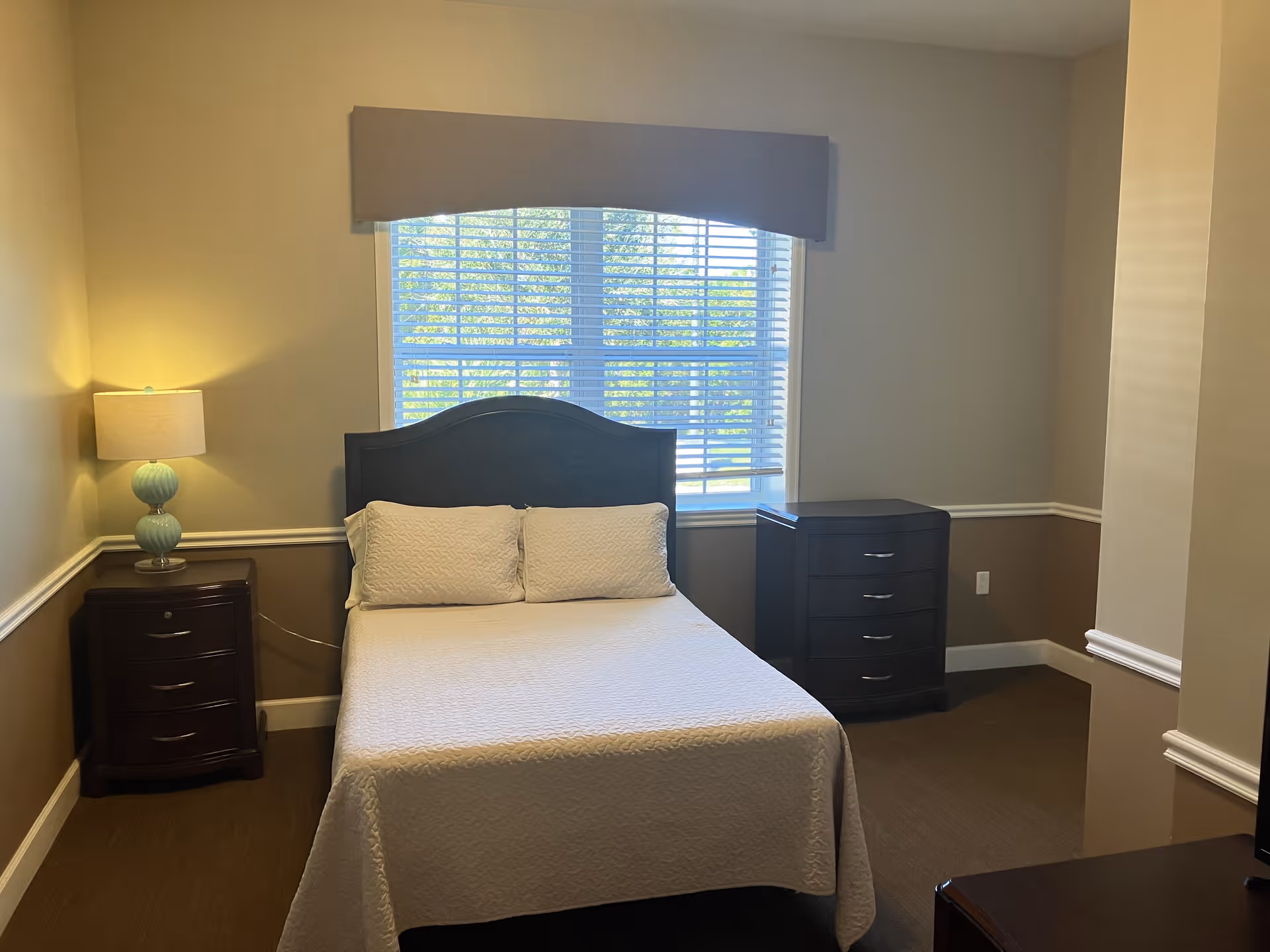 A neatly made bed with white bedding and two pillows is centered against a wall with a window above it. On the left side of the bed is a nightstand with a lamp, and on the right side is a chest of drawers. The room has beige walls with white trim and a brown carpeted floor.