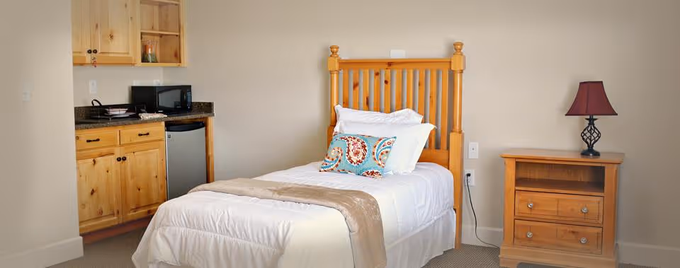A bright assisted-living bedroom with a single bed, wooden headboard, bedside table and a small kitchenette with cabinets and a mini-fridge.