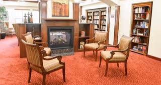 A cozy common area with four wooden armchairs arranged around a fireplace. The room features built-in bookshelves filled with books, a red carpet, and framed artwork above the fireplace. There is a window with curtains on the left side letting in natural light.