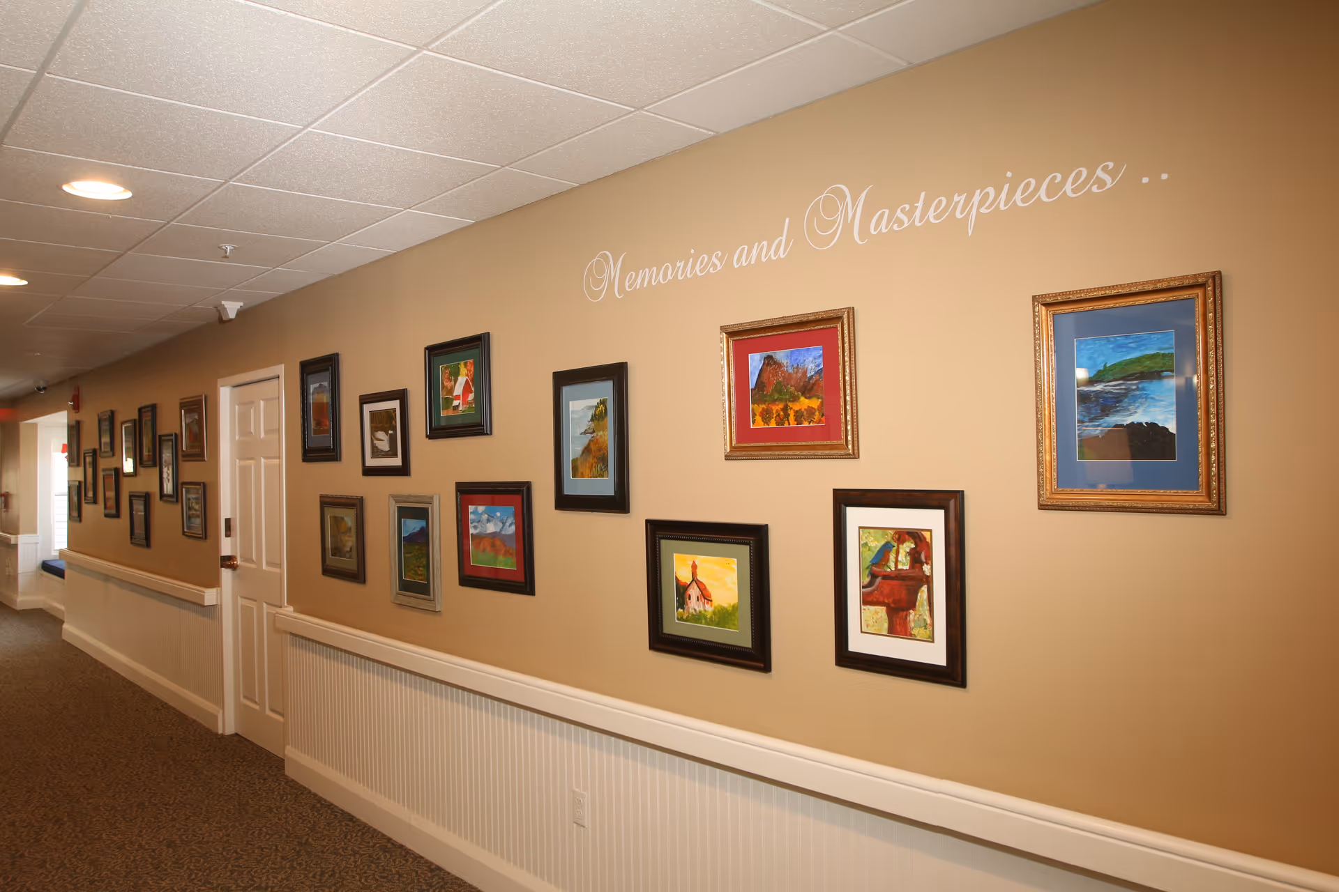 A hallway in a senior living facility with beige walls adorned with multiple framed paintings. Above the paintings, the words 'Memories and Masterpieces...' are written in elegant white script. The hallway has a white door, a white chair rail, and a carpeted floor.
