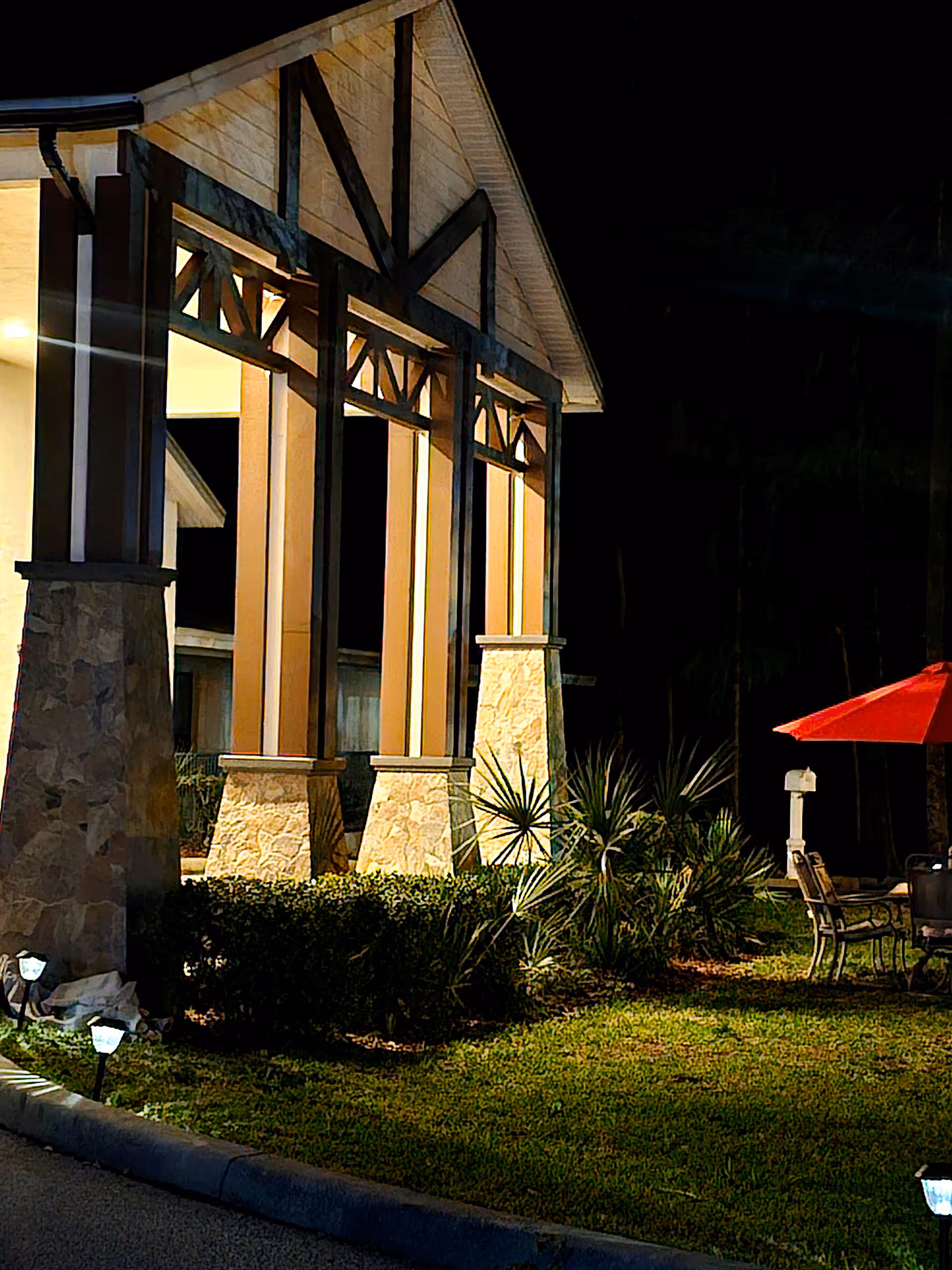 Night view of the exterior entrance area of a building with stone pillars and wooden beams, illuminated by warm lights. There is a small garden with bushes and plants, and outdoor seating with a red umbrella on the right side.
