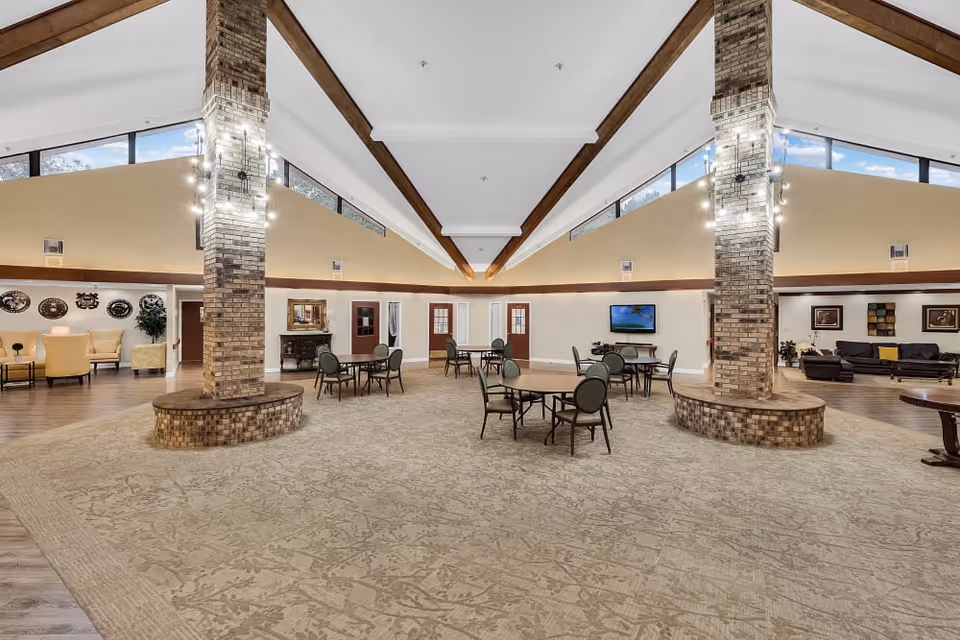 Spacious senior living common area with high vaulted ceilings featuring wooden beams, two large brick columns with modern light fixtures, round tables with chairs arranged on a patterned carpet, and seating areas with sofas and armchairs along the walls under large windows.