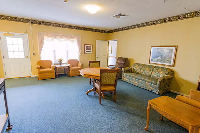 A cozy living room area in Timberdale Trace Assisted Living featuring a green patterned sofa, two orange armchairs with a small table between them near a window with sheer curtains, a wooden table with four chairs in the center, and a framed landscape painting on the wall.
