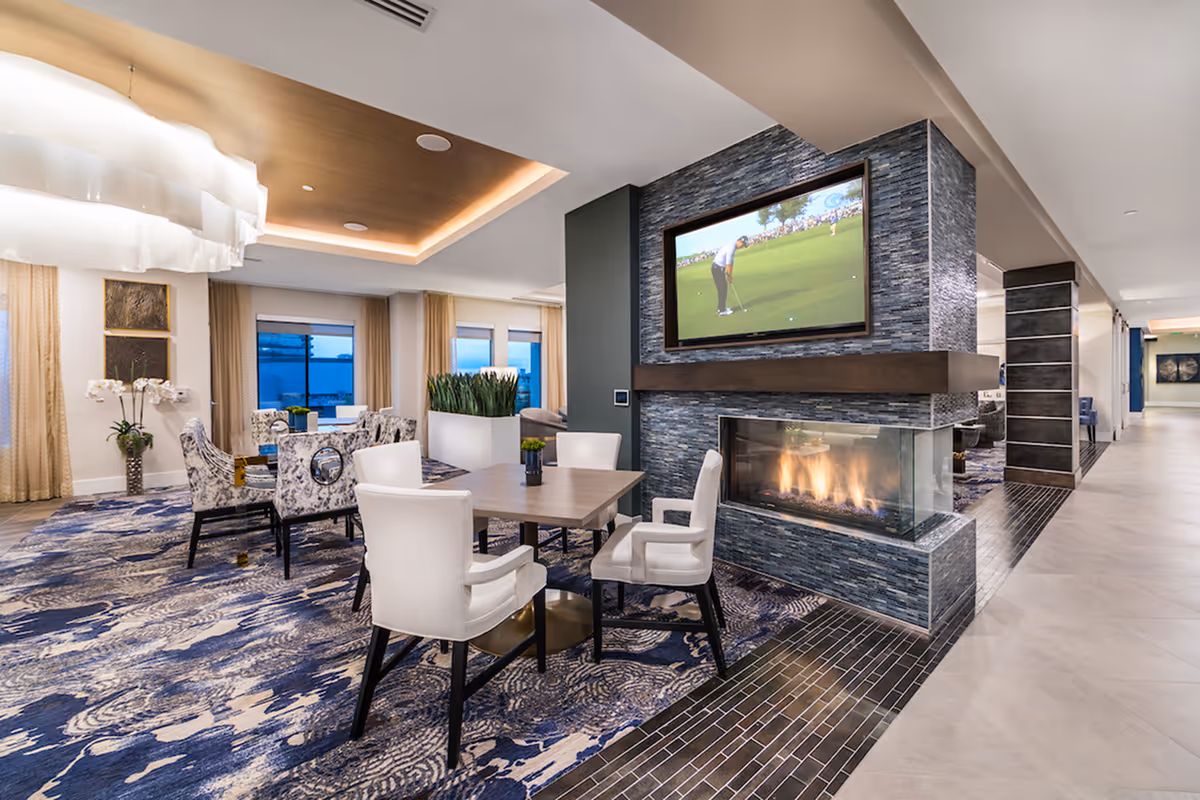 A modern and elegant common area with a cozy fireplace surrounded by blue stone tiles, a flat-screen TV mounted above it showing a golf game, and several seating arrangements including white armchairs and patterned chairs around tables. The room features large windows with beige curtains, decorative plants, and a stylish ceiling with recessed lighting.