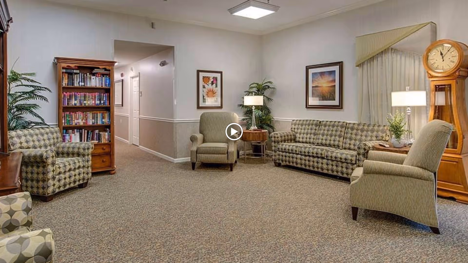 A cozy living room area in Bradford Court featuring patterned armchairs and a matching sofa, a wooden grandfather clock, a bookshelf filled with books, framed artwork on the walls, table lamps, and a potted plant. The room has neutral-colored walls and carpeted flooring.
