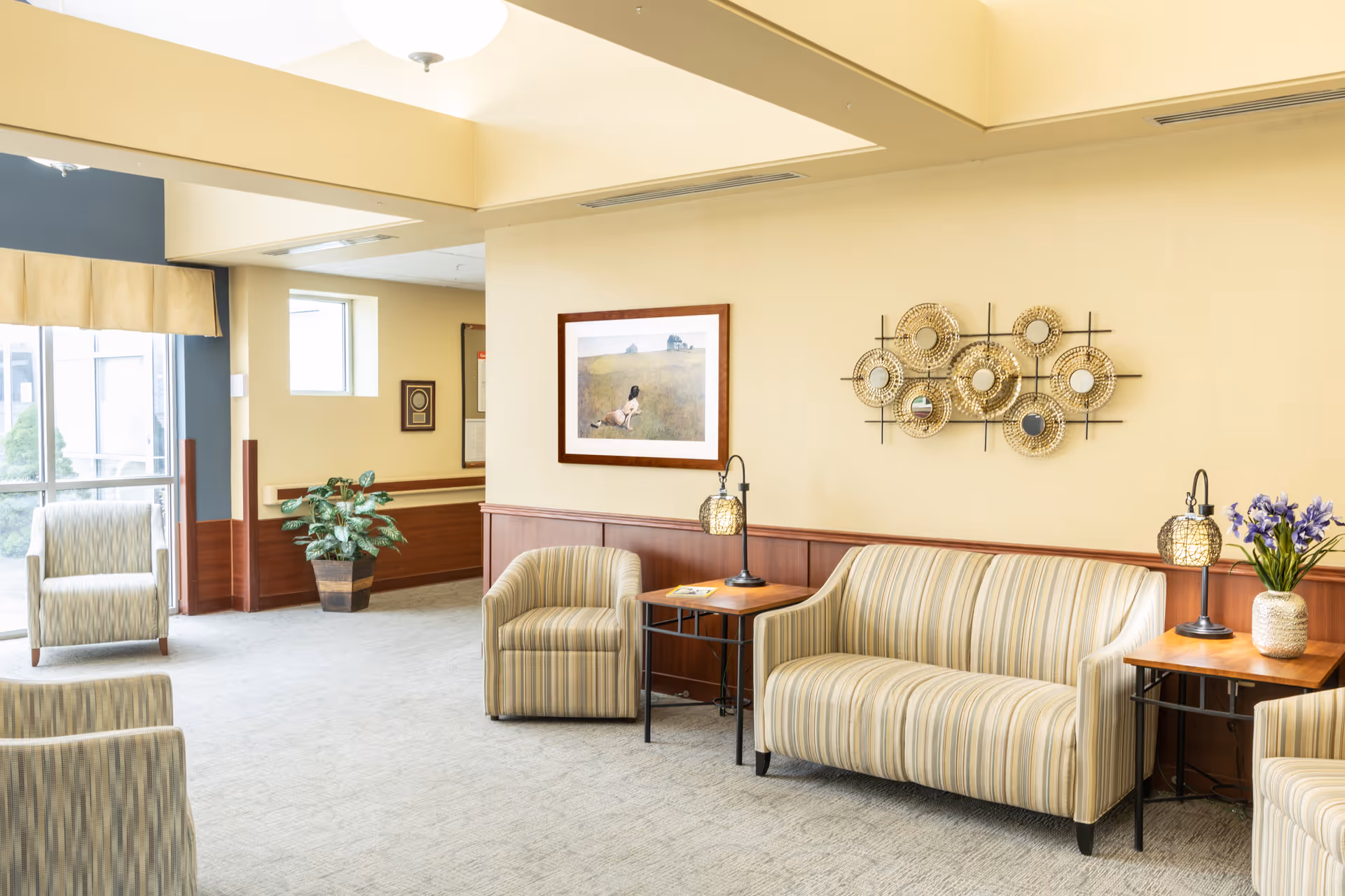 A bright and welcoming sitting area in a senior living facility with striped upholstered chairs and a sofa, two wooden side tables with decorative lamps, a potted plant, wall art including a framed picture and a decorative mirror arrangement, and large windows letting in natural light.