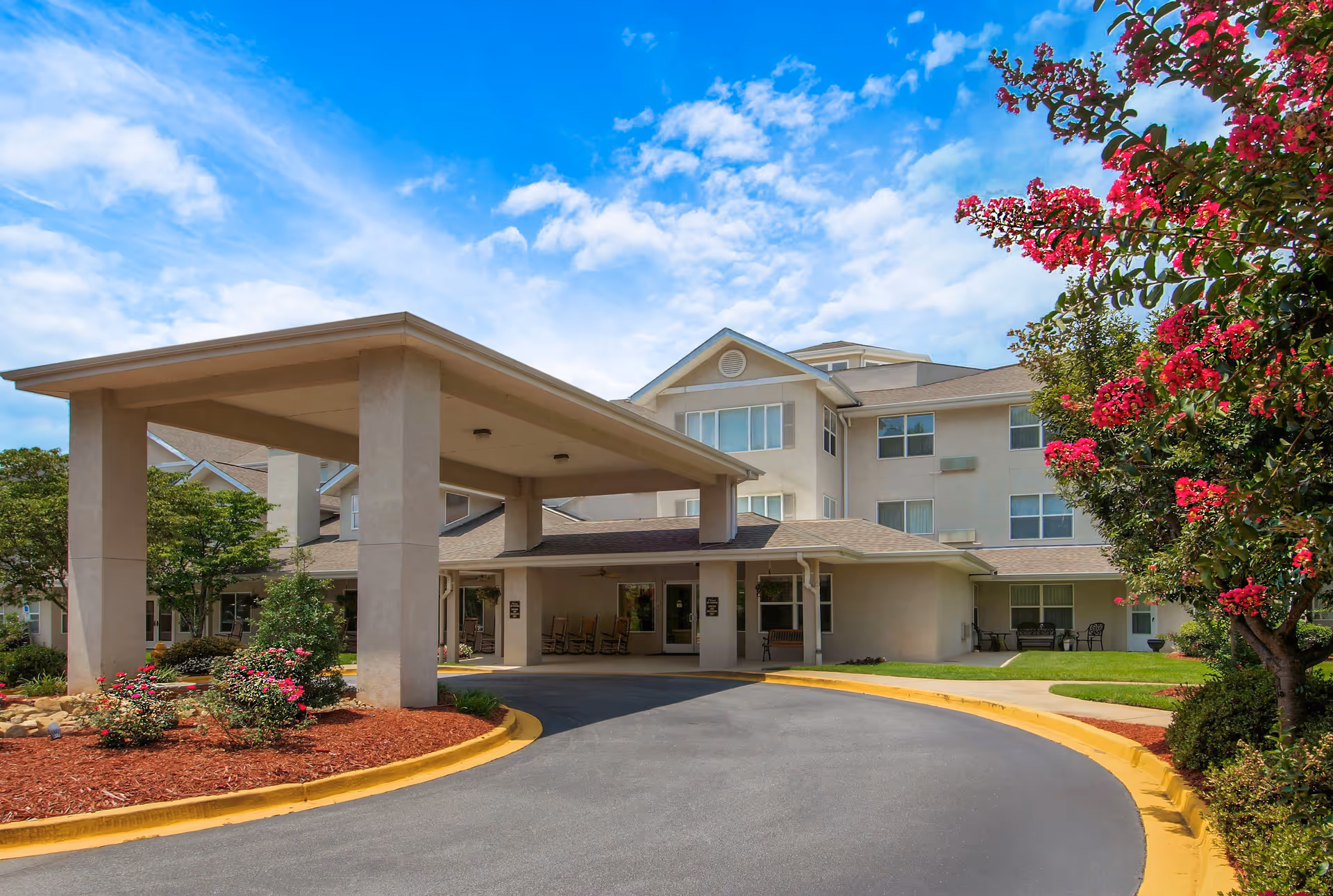 Exterior view of TerraBella Greenville senior living facility showing the main entrance with a covered drop-off area, surrounded by landscaped gardens with flowering bushes and trees under a partly cloudy blue sky.