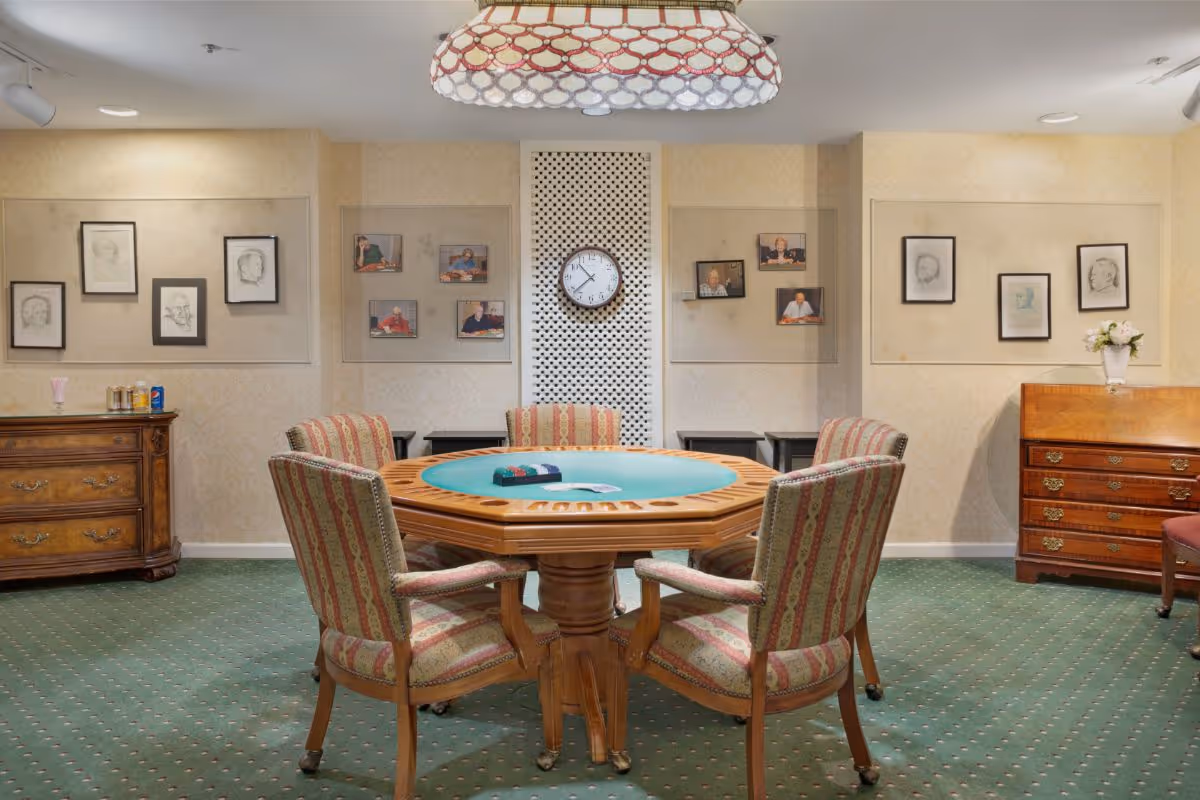A cozy room with a round wooden table designed for card games, surrounded by six upholstered chairs with striped fabric. The room has green carpet with a dotted pattern, beige wallpaper, and framed sketches and photographs on the walls. A clock is mounted on a white lattice panel in the center of the back wall. There are two wooden dressers on either side of the room, one with a vase of flowers and the other with some cans and cups. A decorative hanging light fixture is above the table.