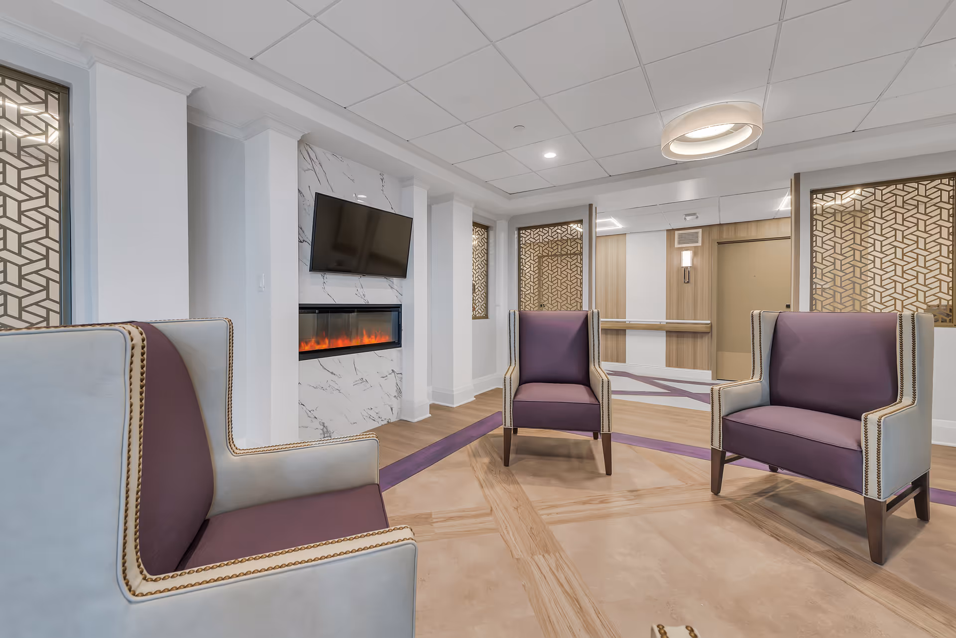 A modern assisted-living lounge with purple upholstered armchairs arranged around a wall-mounted TV above an electric fireplace.