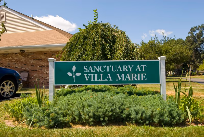 A green sign with white text that reads 'Sanctuary at Villa Marie' is displayed outdoors in front of a bush and some greenery. Behind the sign, there is a brick building with a beige roof and a parked blue car partially visible on the left side. The sky is clear and blue.