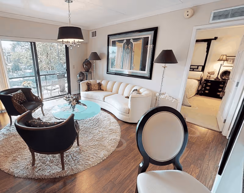 A cozy living room with a curved white sofa, two dark armchairs, a round glass coffee table on a circular beige rug, and a large framed artwork on the wall. There are two tall floor lamps on either side of the sofa. A sliding glass door opens to an outdoor patio with chairs and greenery visible outside. A doorway leads to a bedroom with a bed and nightstand visible.