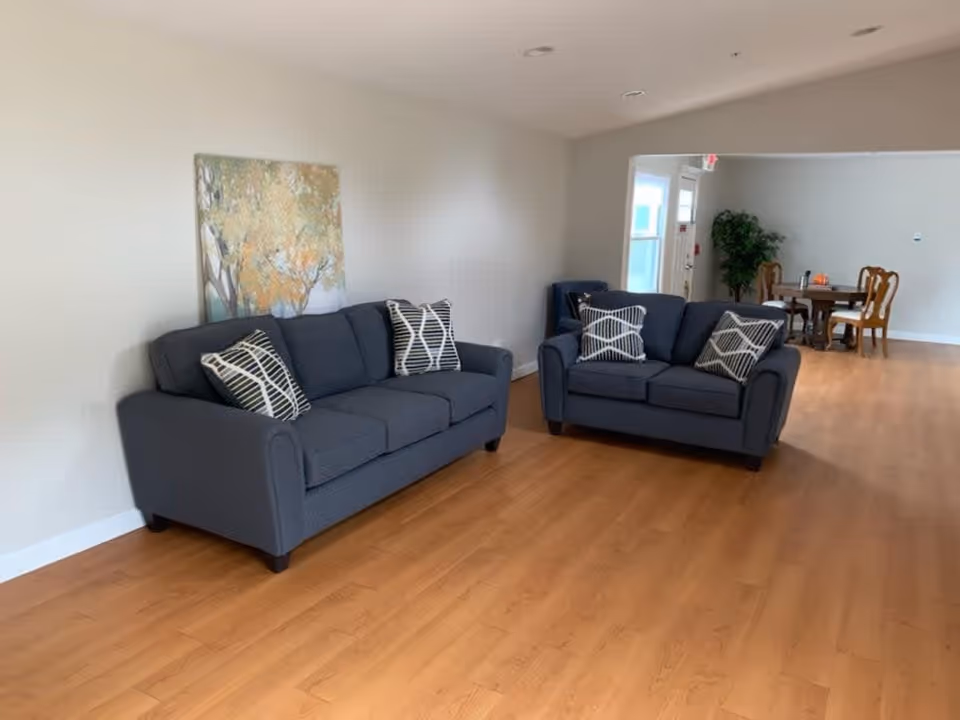 A spacious living area with two dark gray sofas adorned with patterned throw pillows. A large painting with autumn trees hangs on the wall behind one sofa. The room has light wood flooring and white walls. In the background, there is a dining area with a wooden table and chairs, a potted plant, and a door with windows.