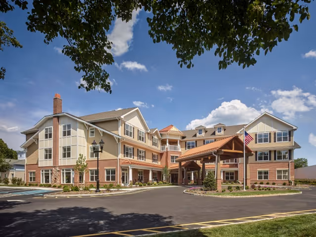Exterior view of a multi-story senior living facility building with a covered entrance, landscaped grounds, and an American flag on a flagpole under a partly cloudy blue sky.