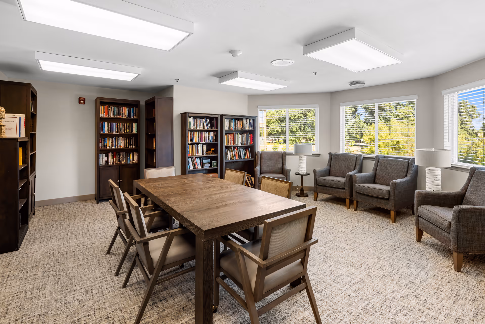 A bright and spacious room with a large wooden table surrounded by eight chairs in the center. Along the walls are several bookshelves filled with books. There are five gray upholstered armchairs arranged near windows that let in natural light, with two small side tables each holding a lamp. The room has a neutral color palette with beige carpet and white walls, creating a cozy and inviting atmosphere.