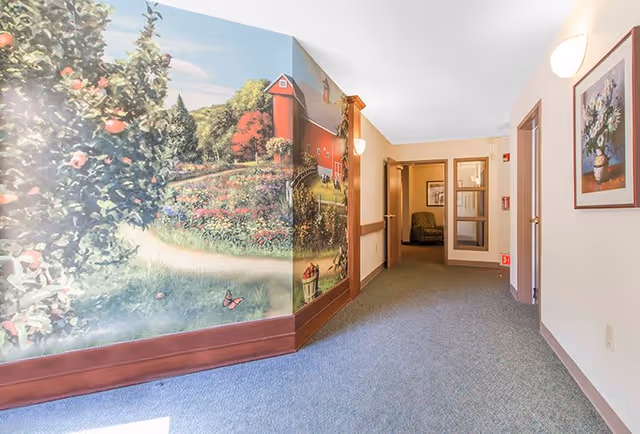 A hallway in a senior living facility with a large mural on the left wall depicting a scenic farm landscape with trees, flowers, and a red barn. The hallway has carpeted flooring, beige walls with wooden trim, a framed floral painting on the right wall, and a doorway leading to a room with a green armchair visible inside.
