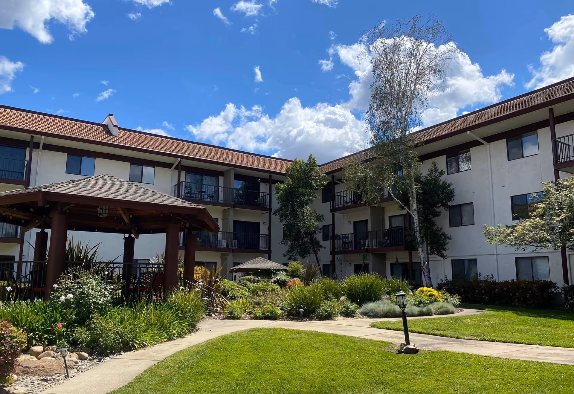 Three-story senior living building surrounding a landscaped courtyard with a gazebo under a blue sky.