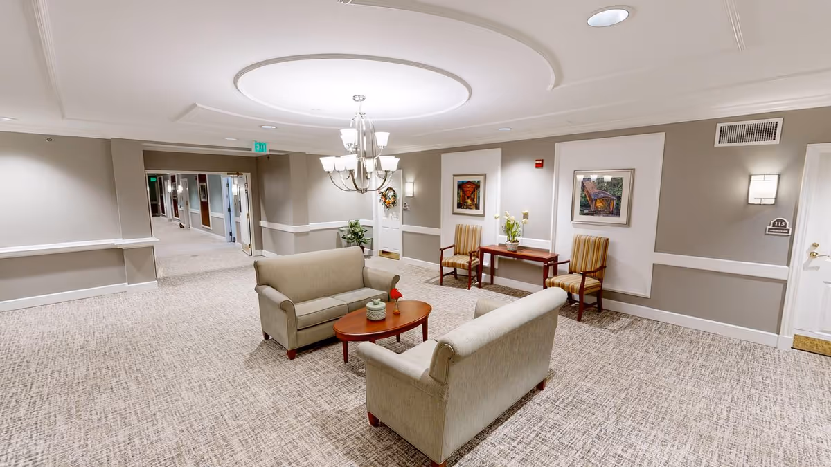 A well-lit common area in a senior living facility featuring two beige sofas facing each other with a wooden coffee table in between. The room has neutral-colored walls with white trim, two framed paintings, two striped armchairs, a small wooden table with a plant, and a chandelier hanging from a circular recessed ceiling light. A hallway extends from the left side of the room.