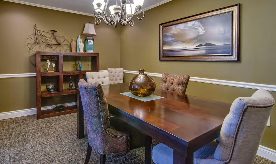A dining room with a dark wooden table surrounded by six upholstered chairs in various patterns and colors. On the table is a large amber glass vase placed on a small blue mat. The walls are painted olive green with white trim, and a framed landscape painting hangs on the wall. A wooden shelving unit with decorative items, including a metal bicycle sculpture, vases, and a small horse figurine, is positioned against the wall. A chandelier with multiple lights hangs from the ceiling.