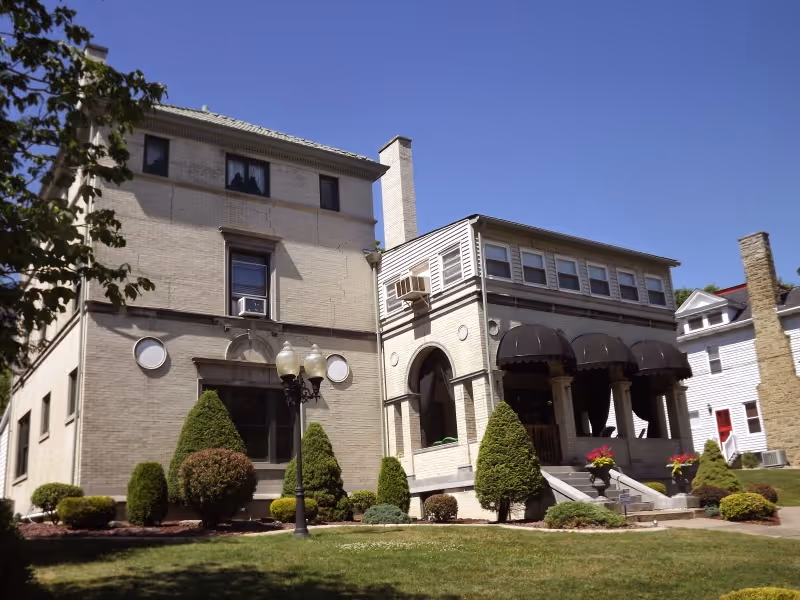 Exterior view of a large, light-colored brick building with multiple windows, a chimney, and a porch with dark awnings. The building is surrounded by neatly trimmed bushes and a well-maintained lawn under a clear blue sky.