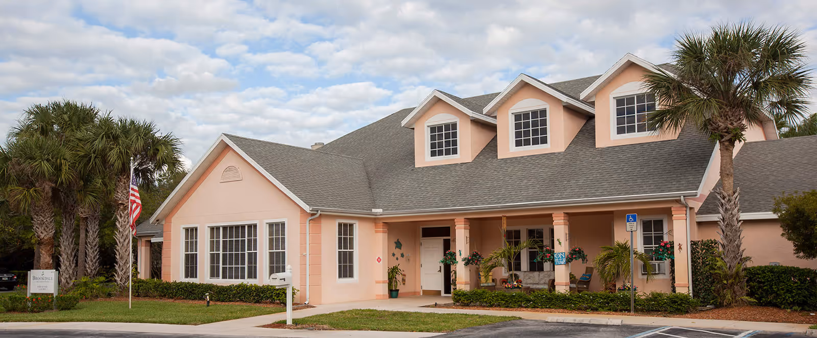 Exterior view of a peach-colored senior living facility building with a gray roof, three dormer windows, palm trees, and a small garden in front. There is a flagpole with an American flag and a sign that reads Brookdale. The entrance has a covered porch with hanging plants and seating.