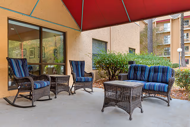 Outdoor patio area with wicker furniture including two rocking chairs, two regular chairs, a loveseat, and two tables. The furniture has blue striped cushions. The patio is covered with a red canopy and is adjacent to a building with beige walls and windows. There are bushes and trees visible in the background.