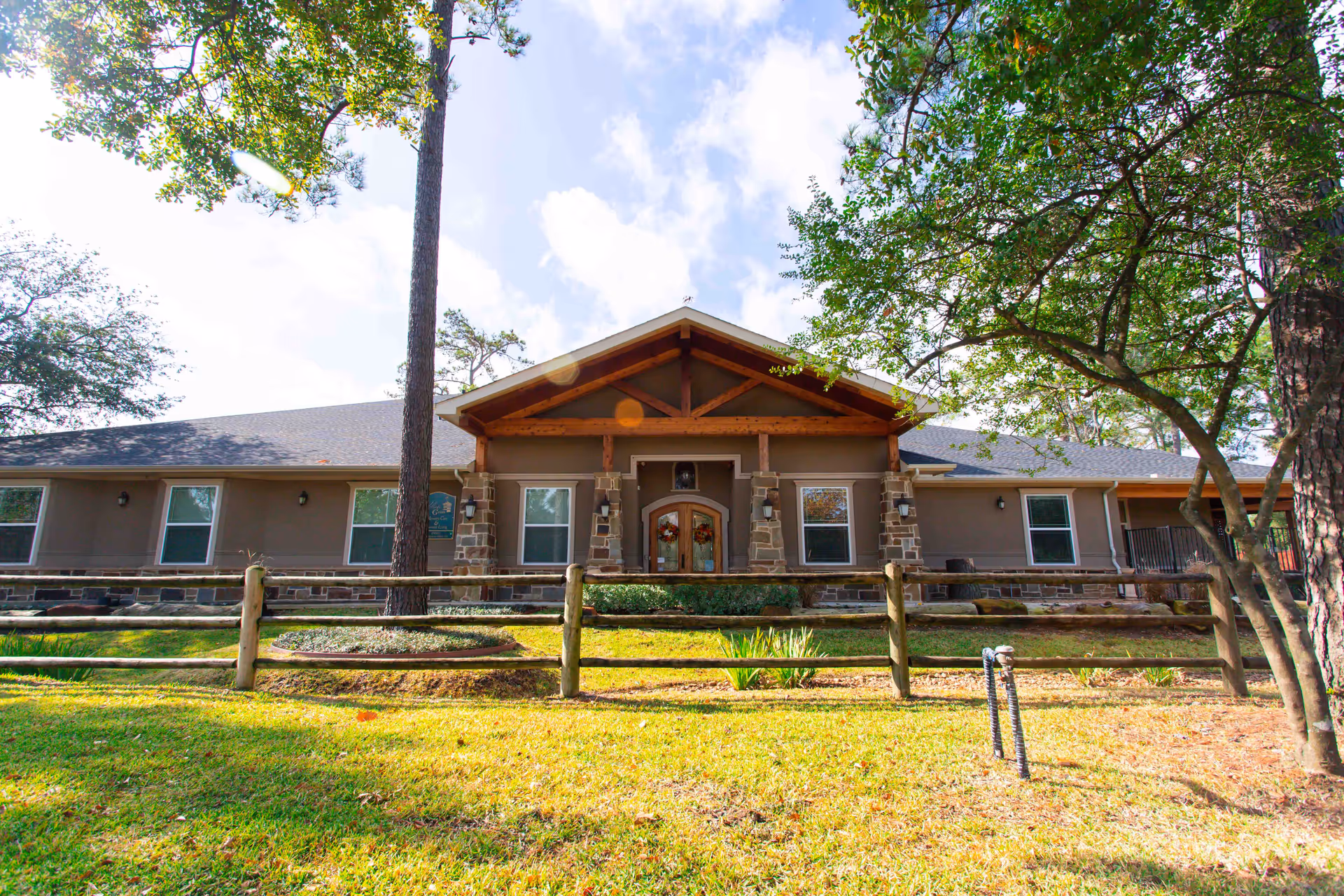 Front exterior view of a single-story building with a peaked roof and stone pillars at the entrance. The building is surrounded by trees and a wooden fence with a grassy lawn in front under a partly cloudy sky.