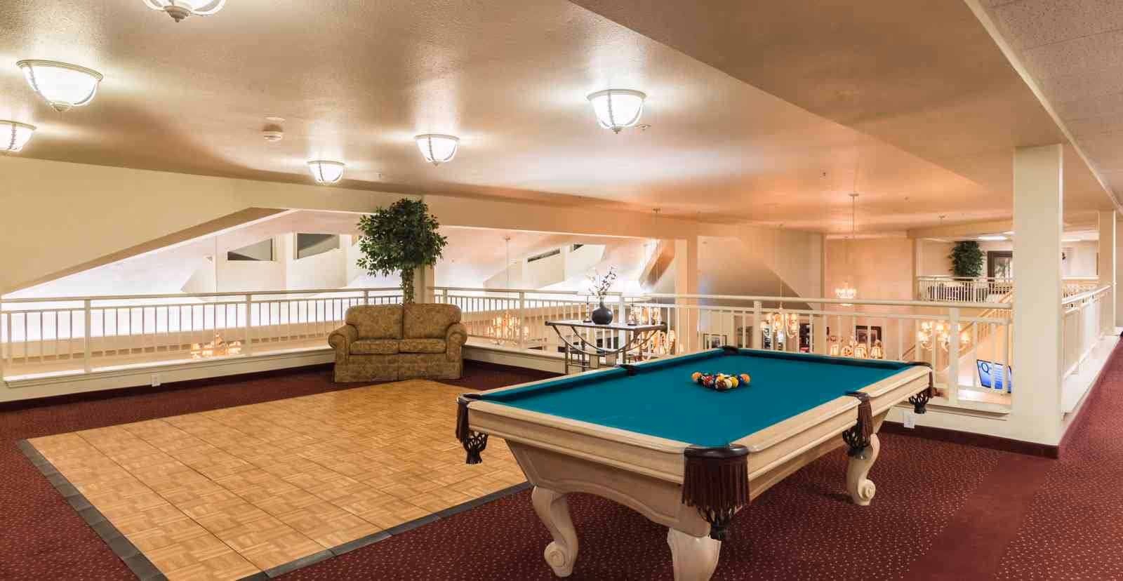 Interior view of a senior living facility's recreational area featuring a pool table with balls arranged for a game, a small table with chairs, a couch, and decorative plants. The space has carpeted floors with a wooden parquet section, ceiling lights, and white railings overlooking a lower level with chandeliers.
