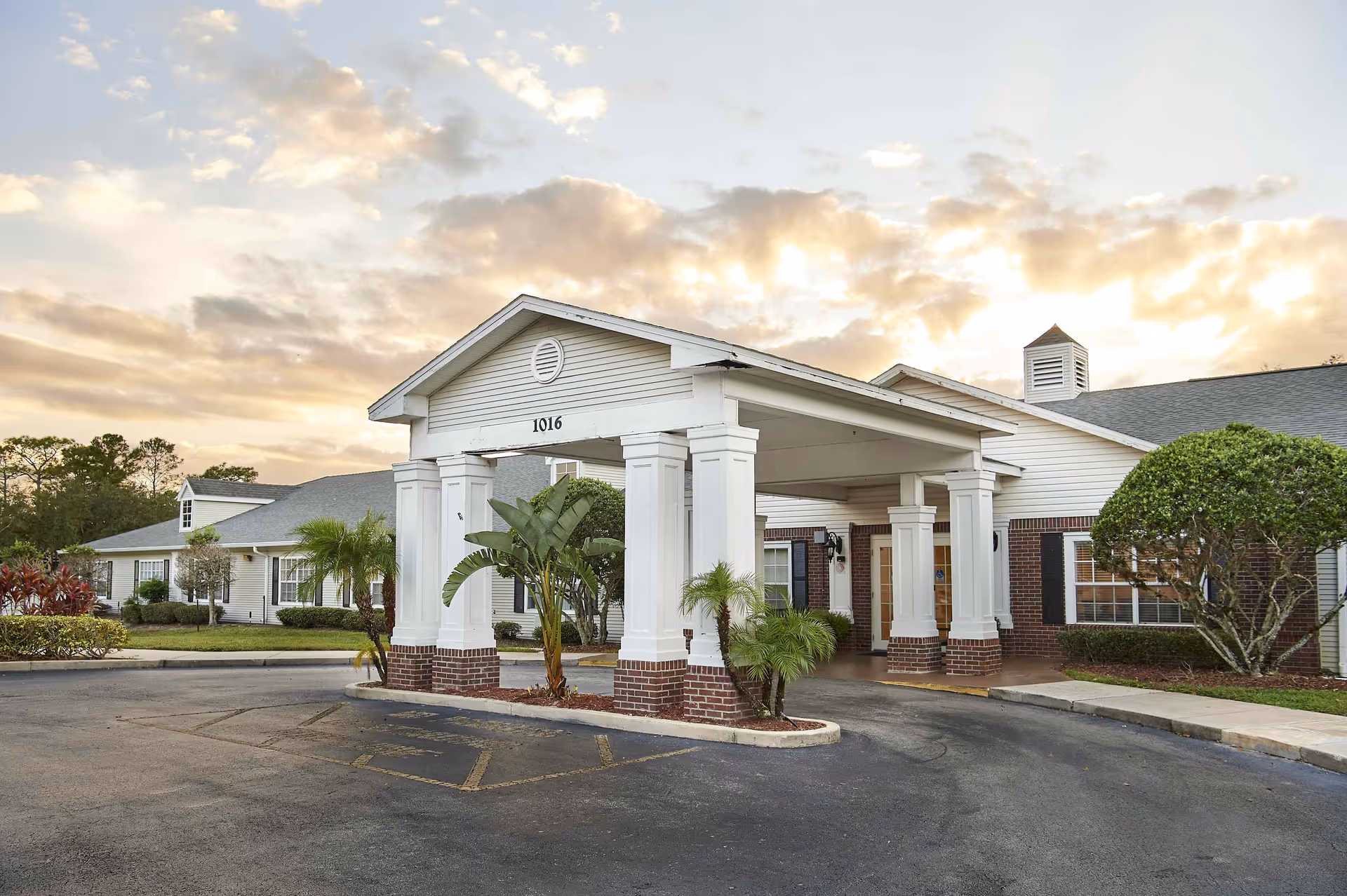 Exterior view of Palmetto Landing senior living facility showing a covered entrance with white columns and brick bases, surrounded by landscaped greenery and a partly cloudy sky at sunset.