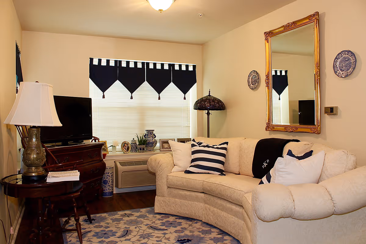 Cozy living room with a cream curved sofa and striped pillows, a TV on a wooden table, decorative mirror and window valance.
