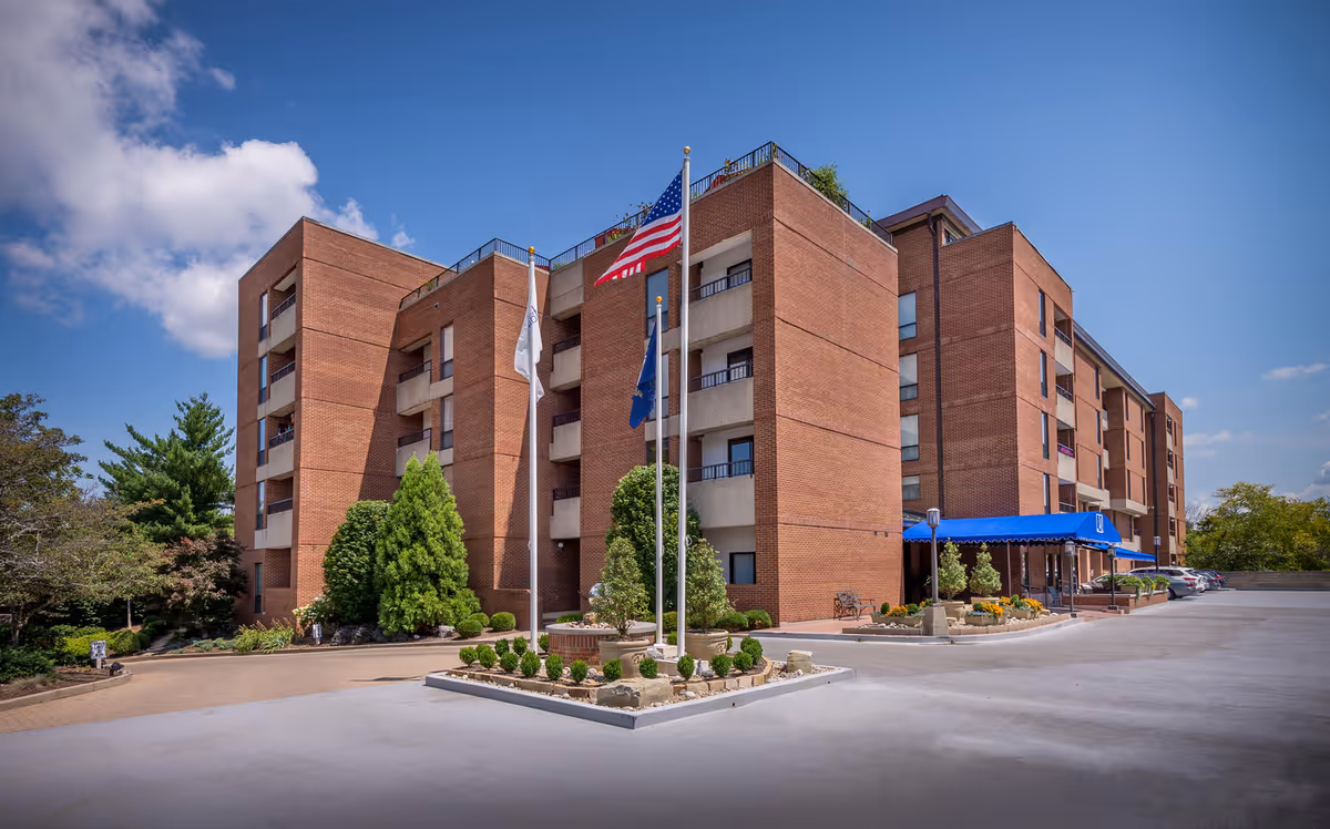 Exterior view of a multi-story brick building with balconies, three flagpoles with flags, landscaped greenery, and a blue awning over the entrance on a sunny day with a partly cloudy sky.