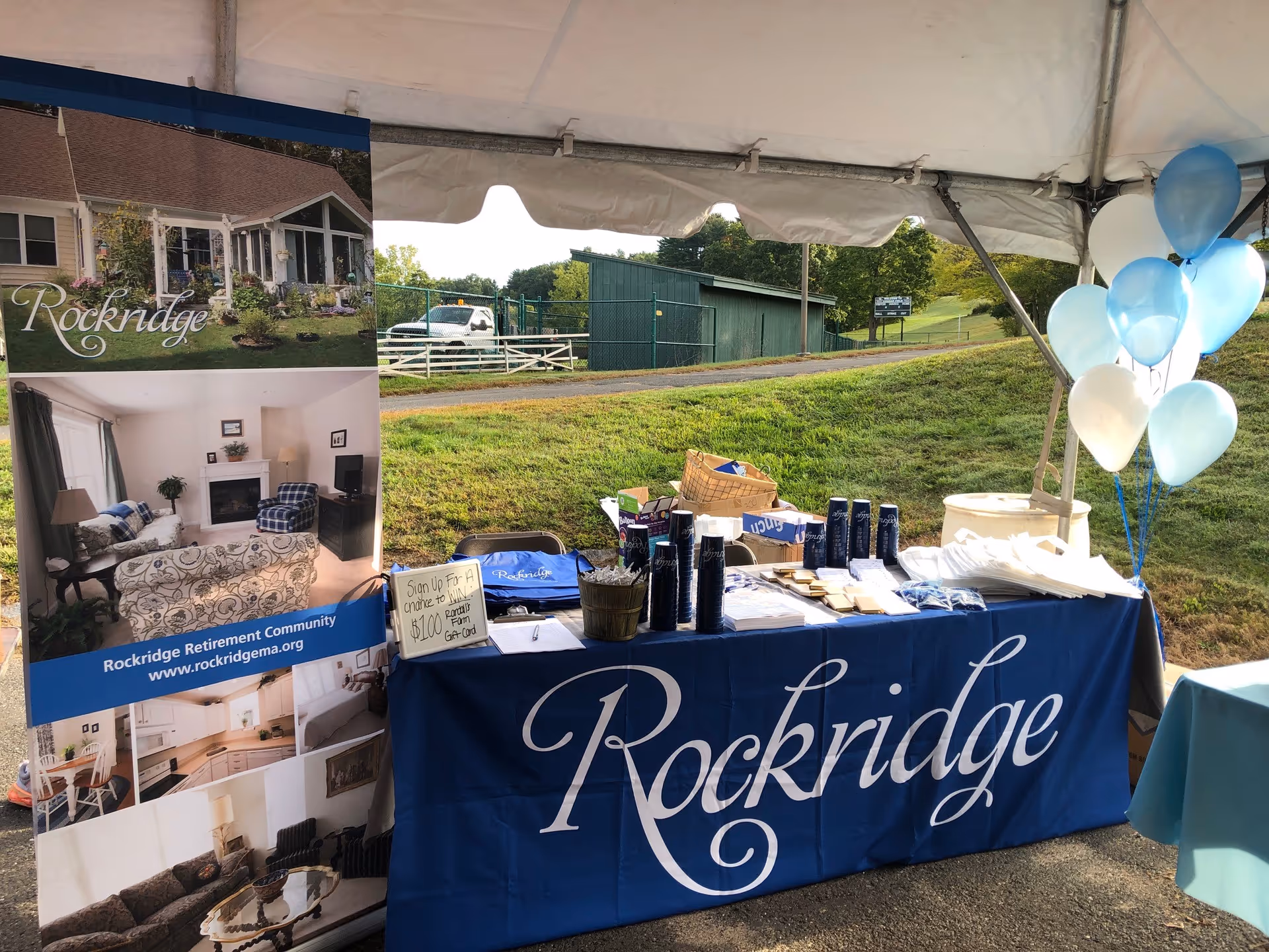 Outdoor promotional booth for Rockridge Retirement Community under a white tent, featuring a blue tablecloth with the Rockridge logo, informational brochures, giveaway items, a sign about a $10 raffle, and blue and white balloons. A banner with images of the community's exterior and interior is displayed to the left.