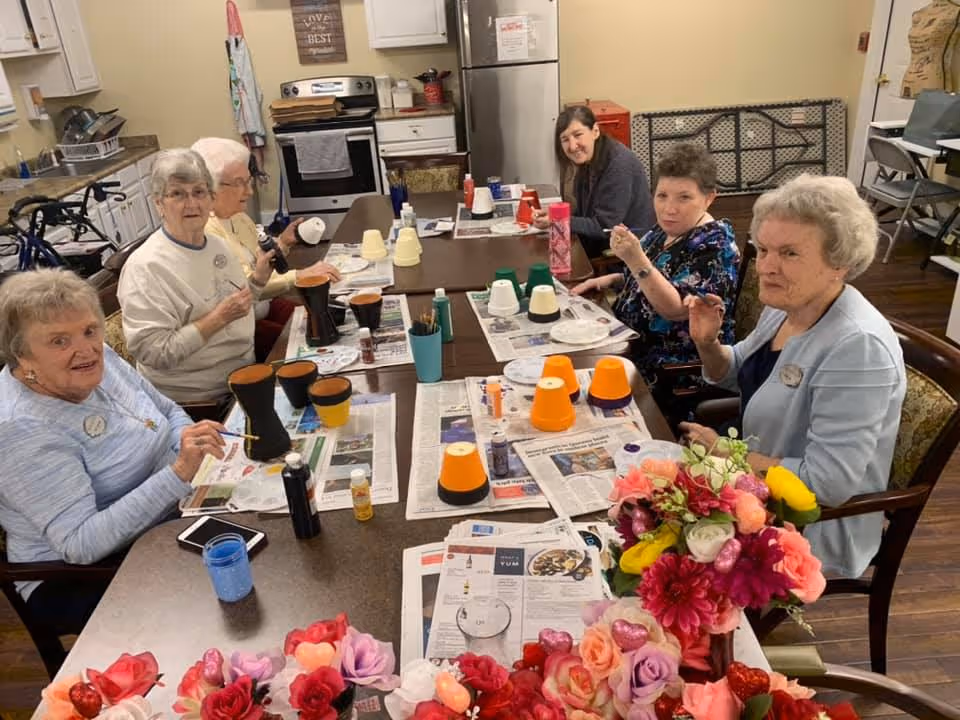 A group of elderly women sitting around a table in a kitchen or craft room, painting small flower pots. The table is covered with newspapers and various painting supplies. There are colorful artificial flowers in the foreground.