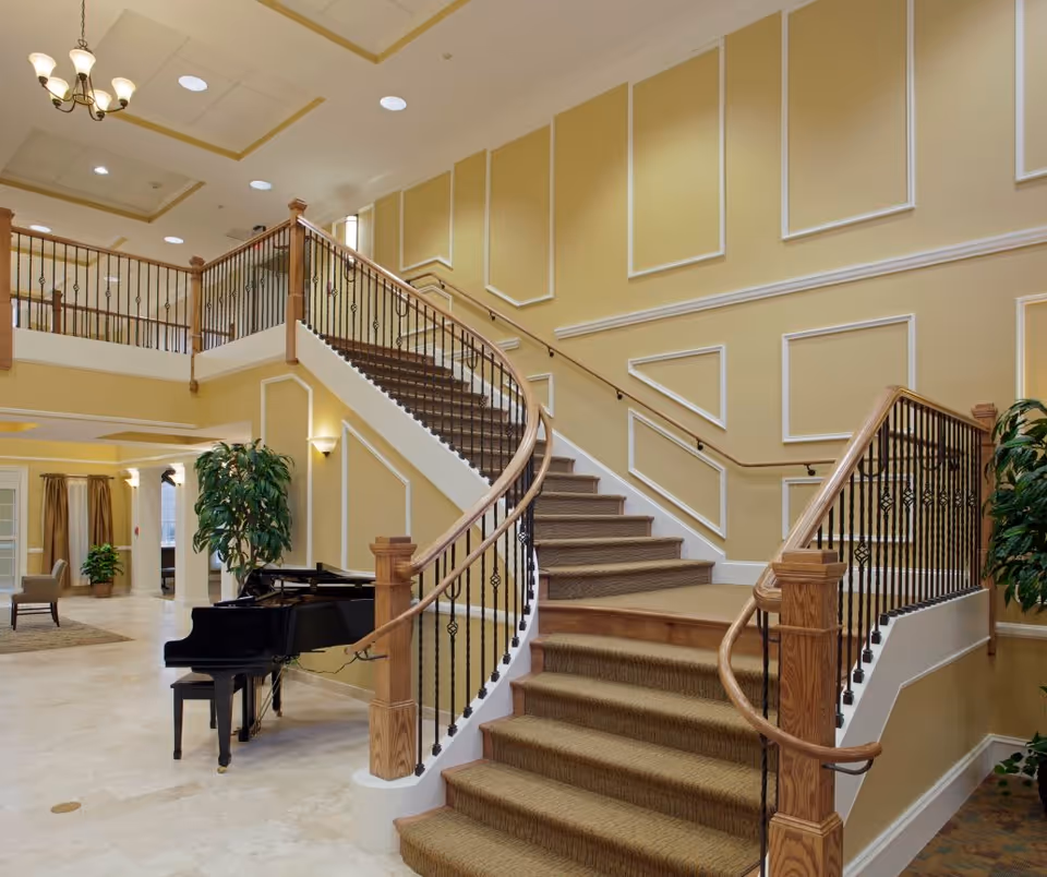 Interior view of a senior living facility featuring a grand curved staircase with wooden handrails and black iron balusters. The walls are painted yellow with white decorative molding. A black grand piano with a matching bench is placed near the base of the stairs. There are large potted plants and seating areas visible in the background under warm lighting.