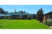 Single-story brick retirement building with a wide green lawn, flagpole, and landscaped shrubs under a clear sky.