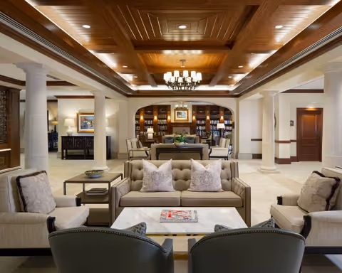 A spacious and elegant living room area with a wooden coffered ceiling, beige tiled floor, and white columns. The room features a beige tufted sofa, matching armchairs, a marble coffee table with magazines, and a bookshelf with decorative items and framed artwork in the background.