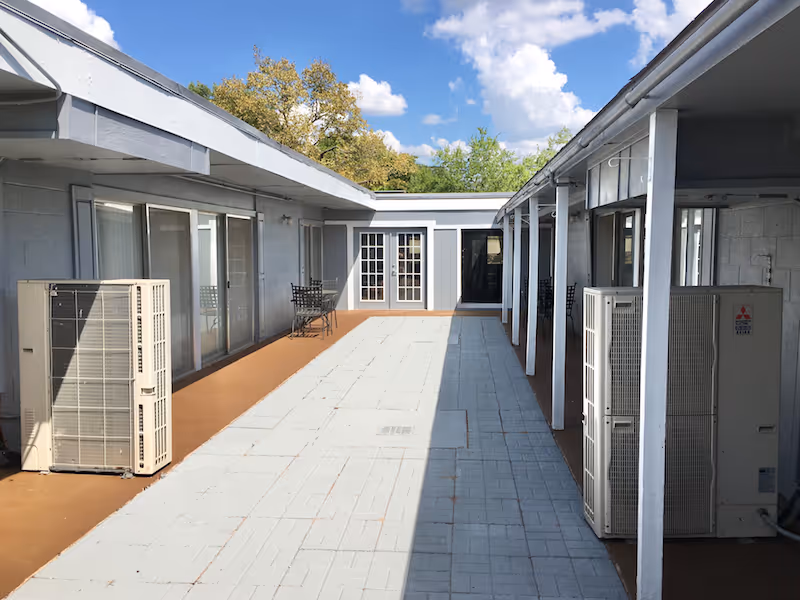 Outdoor courtyard area at Exbury Garden Assisted Living Community with a paved walkway in the center, flanked by building walls with windows and doors. There are two outdoor air conditioning units on either side and a small table with chairs on the left side. Trees and a partly cloudy sky are visible in the background.