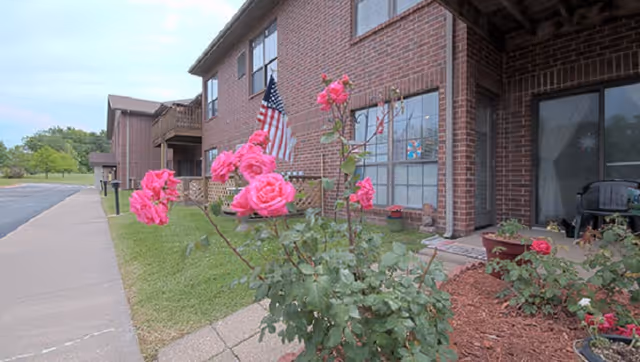 Brick apartment building exterior with pink roses in the foreground, a sidewalk, and an American flag by a ground-floor patio.