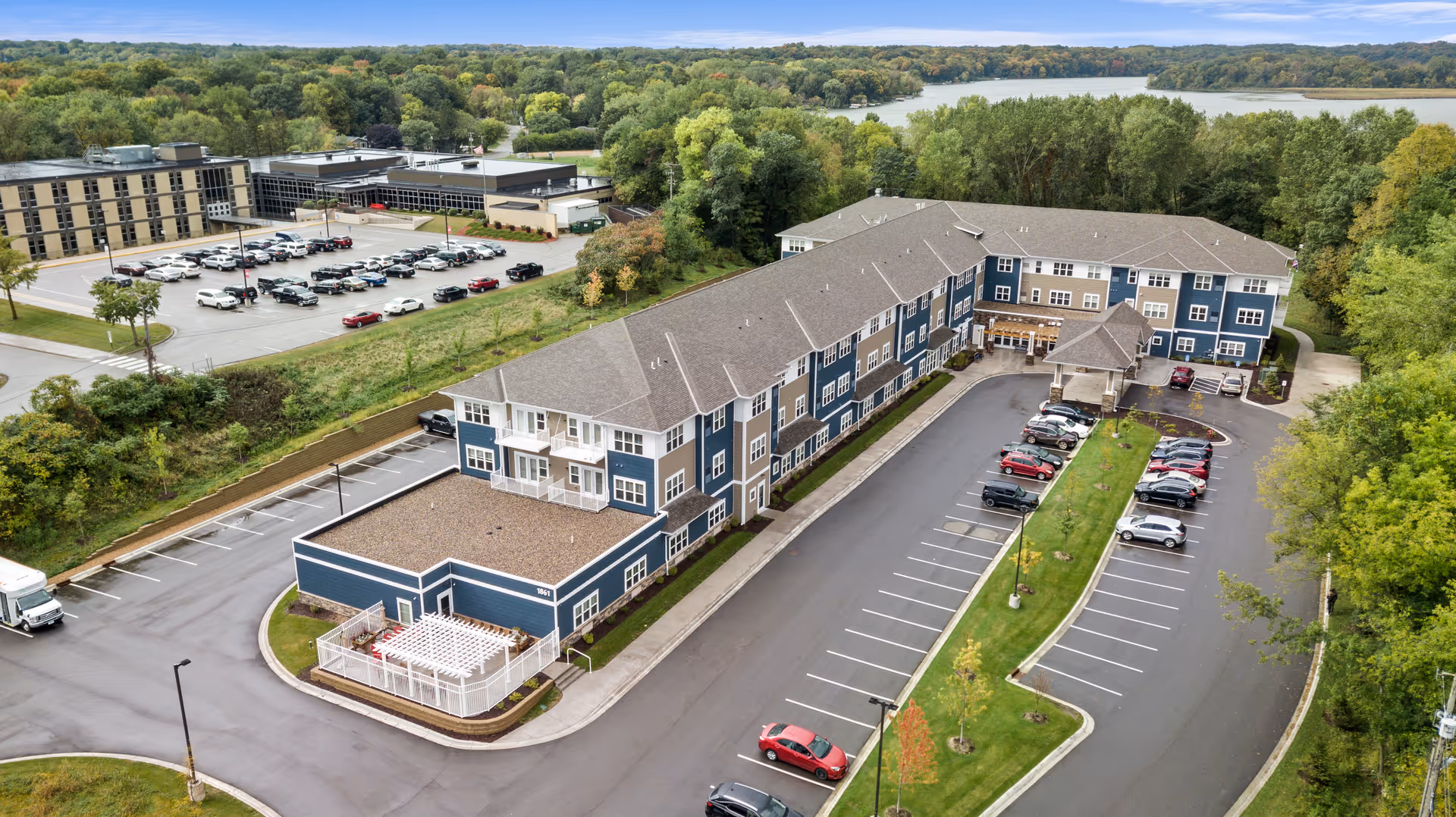 Aerial view of Harrison Bay Senior Living facility showing a large three-story building with blue and beige exterior, surrounded by parking lots with cars, greenery, and trees. In the background, there is a lake and forested area under a clear sky.