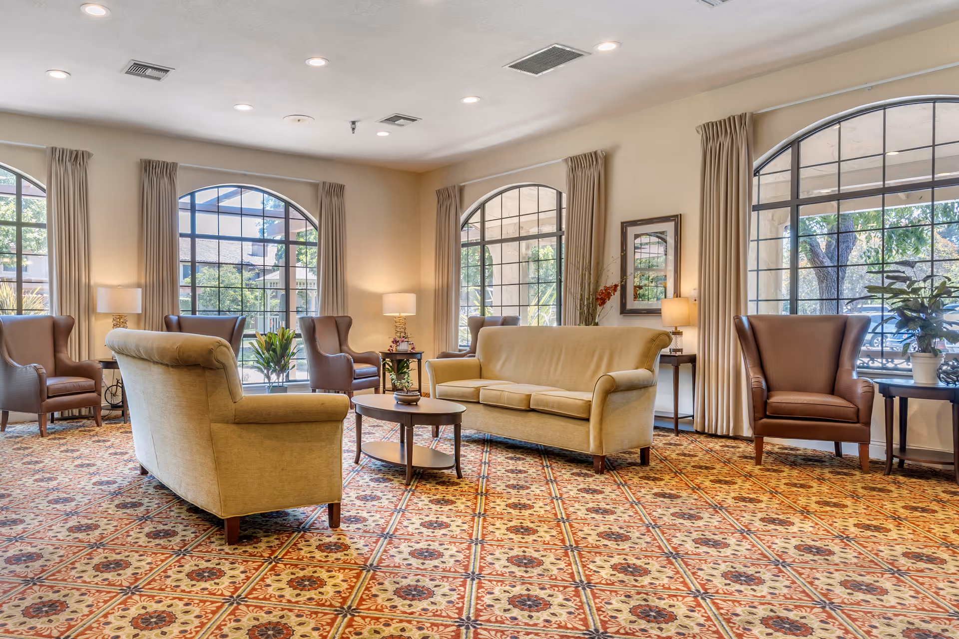 A spacious and well-lit living room area with large arched windows letting in natural light. The room features beige sofas, brown armchairs, several side tables with lamps, and decorative plants. The floor is covered with a patterned carpet in warm tones.