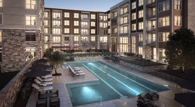A rectangular outdoor swimming pool in a courtyard surrounded by a multi-story residential building with lounge chairs and umbrellas at dusk.
