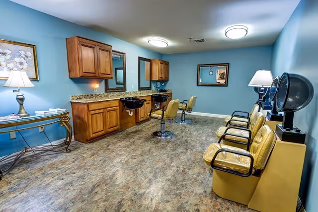 Interior of a hair salon with blue walls, two black shampoo sinks with yellow salon chairs, a row of yellow waiting chairs with hair dryers, wooden cabinets, a decorative table with a lamp and framed artwork on the wall.