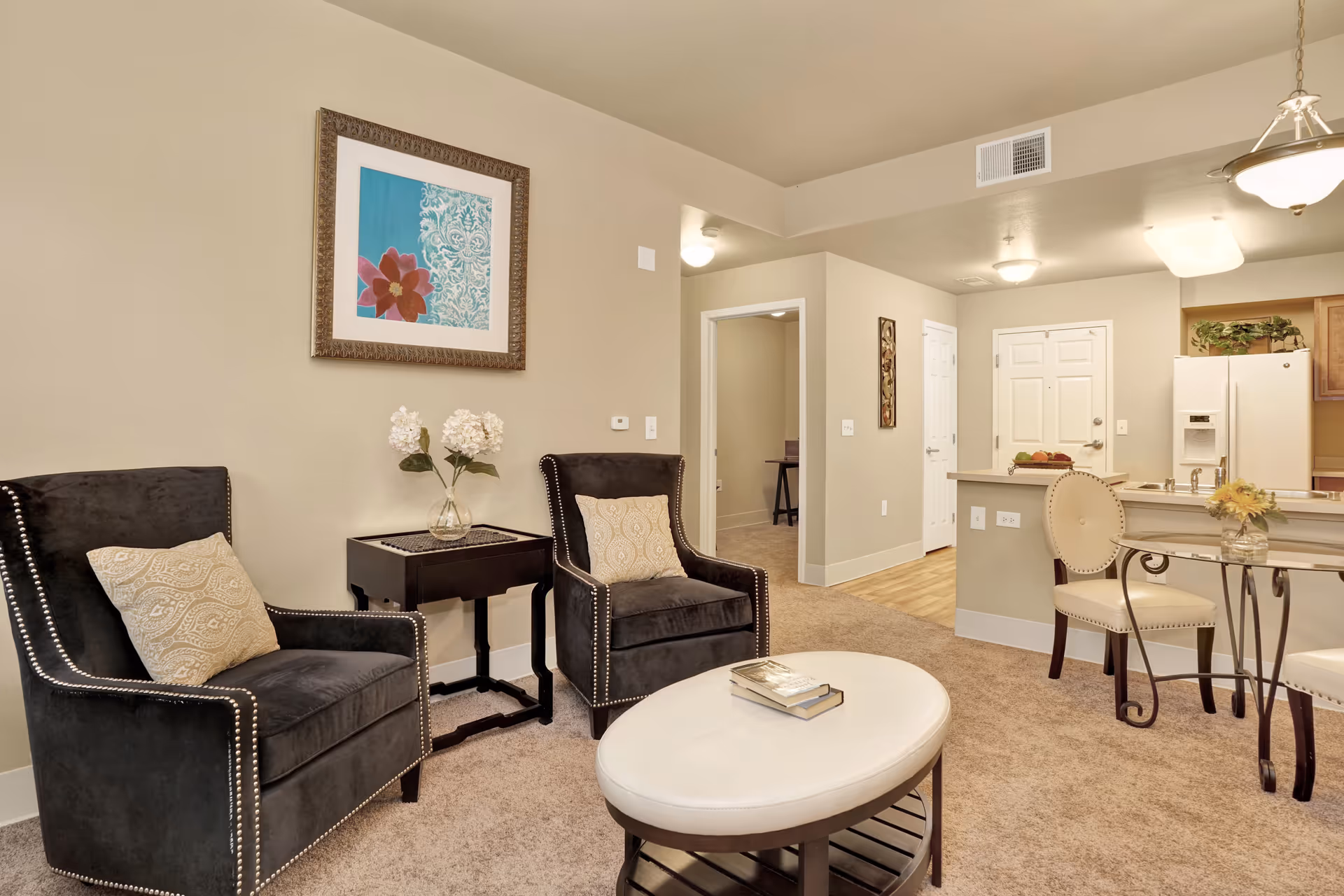 A cozy living room area with two black armchairs with beige patterned pillows, a small dark wooden side table with a vase of white flowers, and a round white coffee table with books on it. The room has beige walls, a framed artwork with a blue and red floral design, and an open view into a kitchen with a white refrigerator, wooden cabinets, and a breakfast bar with two cream-colored chairs. The floor is carpeted in the living area and transitions to wood in the kitchen.